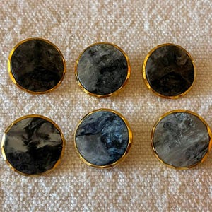 Può includere: Un set di sei bottoni circolari caratterizzati da un motivo marmorizzato nei toni del nero, grigio e bianco. Ogni bottone è racchiuso in un bordo in metallo color oro lucido con un profilo sfaccettato, creando un contrasto tra centro e cornice.