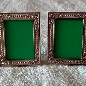 Pode incluir: Dois porta-retratos retangulares com uma borda metálica em tom de bronze, apresentando um intrincado padrão de volutas em relevo e uma borda interna com contas. Cada moldura contém uma inserção verde vibrante e sólida no centro.