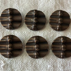 May include: Six bronze-colored, round buttons with a textured design. Each button features three horizontal ridges and a vertical line of small, circular details. The buttons are arranged on a white, textured surface.