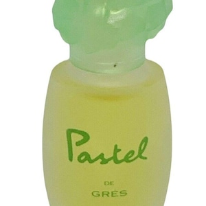 Pode incluir: Um frasco de perfume vintage "Pastel" da Grès. O frasco tem uma tampa verde clara em forma de flor e um corpo amarelo pálido. As palavras "Pastel", "DE GRES" e "EAU DE TOILETTE" são impressas no frasco.