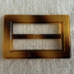 Puede incluir: Hebilla rectangular con un patrón de carey. La hebilla está hecha de un material translúcido con tonos marrones y ámbar. Tiene una abertura rectangular en el centro y una barra en el medio.