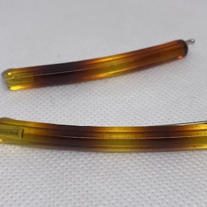 Könnte beinhalten: Zwei Haarklammern mit Schildpattmuster. Die Klammern sind gebogen und weisen einen Farbverlauf von Gelb nach Braun auf. Jede Klammer hat einen silberfarbenen Metallstift an einem Ende. Auf weißem Untergrund.