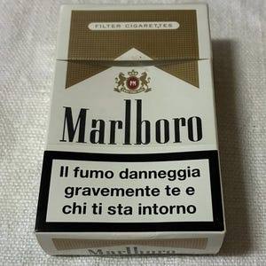 May include: A white and gold Marlboro cigarette pack. The pack features the Marlboro logo in bold black letters, a warning label in Italian, and the text "FILTER CIGARETTES" at the top.