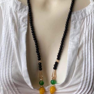 Può includere: Una lunga collana con perline nere, con una sezione di perline gialle, rosse e verdi. La collana ha un grande pendente giallo a forma di goccia. La collana è mostrata su una camicia bianca.