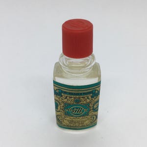 Pode incluir: Uma pequena garrafa de vidro transparente de colônia 4711 Echt Kölnisch Wasser com tampa vermelha. O rótulo apresenta desenhos e texto ornamentados em verde-azulado e dourado. A garrafa é retangular com bordas arredondadas.
