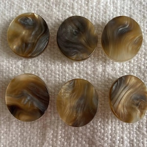 Pode incluir: Seis botões redondos com aparência marmorizada em tons de castanho, bege e cinza. Os botões têm um acabamento brilhante e um padrão em espiral, sugerindo um material natural. Estão dispostos sobre uma superfície branca texturizada.