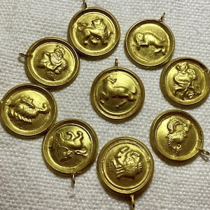 Pode incluir: Uma coleção de pingentes dourados do zodíaco. Cada pingente circular apresenta um símbolo do zodíaco em relevo, incluindo um carneiro, um caranguejo e um leão. Os pingentes têm uma pequena argola na parte superior.