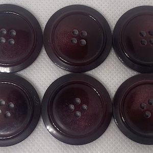 Può includere: Sei grandi bottoni rotondi di colore bordeaux scuro. Ogni bottone ha quattro fori e un bordo esterno leggermente rialzato. I bottoni hanno una lucentezza sottile, che suggerisce una finitura liscia.