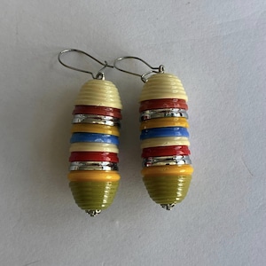 Pode incluir: Um par de brincos pendentes compostos por uma pilha vertical de discos circulares multicoloridos em creme, vermelho, prata, amarelo, azul e verde-oliva. Os brincos estão suspensos em ganchos de metal prateado e têm uma textura canelada.