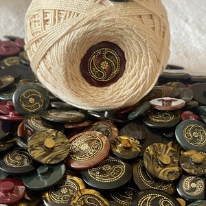 May include: A ball of cream-coloured yarn sits on a pile of vintage buttons. The buttons are in dark shades of black, brown, and green, featuring a gold paisley pattern. Some buttons have a marbled appearance.