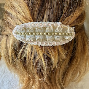 Pode incluir: Uma presilha de cabelo oval com um design de renda branca, acentuada com contas douradas e detalhes em rosa. A presilha está presa ao cabelo castanho claro. Acessório para segurar o cabelo.