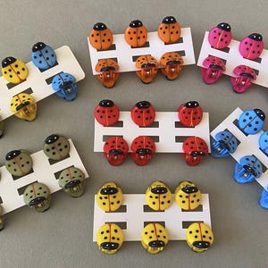 May include: A selection of ladybug-shaped decorative buttons in various colours. The buttons are red, orange, pink, yellow, blue, and green, each with black spots. They are arranged on white card. Metric measurements are not applicable.