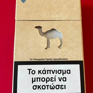 Może przedstawiać: Prostokątne, jasnobrązowe opakowanie papierosów z wyciętą sylwetką wielbłąda na środku. W lewym górnym rogu znajduje się niebieski romb z liczbą 20 i napisem natural flavor. Na dole umieszczono czarno-białą etykietę z ostrzeżeniem zdrowotnym.