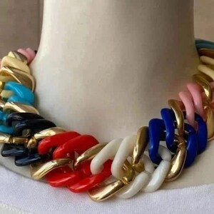 Pode incluir: Um colar arrojado com elos grandes e coloridos. A corrente apresenta uma mistura de cores, incluindo vermelho, branco, dourado, preto, azul, rosa e turquesa. O colar é exibido em um manequim branco.
