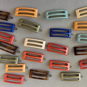Peut inclure: Une collection de barrettes à cheveux rectangulaires de différentes couleurs, dont écaille de tortue, orange, bleu, vert et crème. Chaque barrette présente un design à double fente et un fermoir en métal. Les barrettes sont disposées sur une surface gris clair.