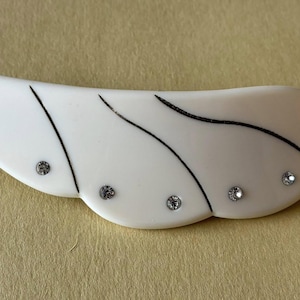 Pode incluir: Grampo de cabelo branco em forma de asa com linhas pretas curvas e strass brilhantes. O grampo tem aproximadamente 10 cm de comprimento e é um acessório de cabelo decorativo.