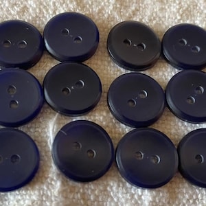 Puede incluir: Doce botones redondos de color azul marino con dos agujeros cada uno, dispuestos en tres filas de cuatro sobre una superficie texturizada de color crema. Ideales para proyectos de costura.