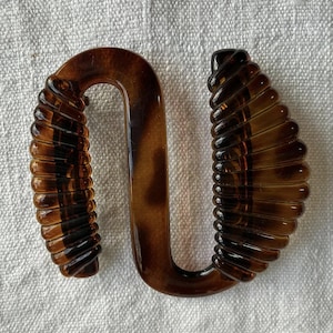 Könnte beinhalten: Eine braune, U-förmige Schnalle mit geripptem, strukturiertem Design auf jeder Seite. Die Schnalle besteht aus einem durchscheinenden Material mit dunkleren braunen Streifen. Der Hintergrund ist ein weißes, strukturiertes Gewebe.