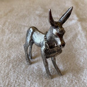 Könnte beinhalten: Silberfarbene Eselfigur mit Kette und Schild "BURRO-TAXI". Der Esel steht auf allen Vieren, die Ohren aufgestellt. Die Figur hat eine detaillierte, strukturierte Oberfläche. Ein dekoratives Objekt.