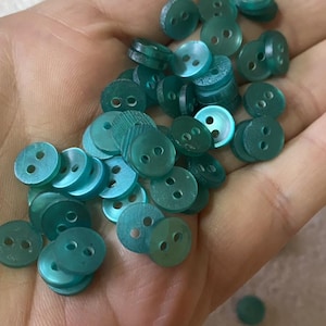 Op de afbeelding: Een verzameling turquoise, ronde knopen met elk twee gaatjes. De knopen zijn verspreid, sommige opgestapeld, en lijken gemaakt van een doorschijnend materiaal. De afbeelding toont een close-up van de knopen.