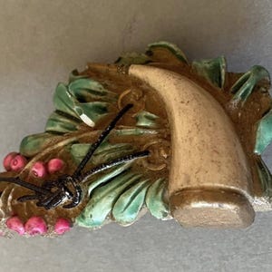 Pode incluir: Broche vintage com design em forma de chifre, folhas verdes, bagas cor-de-rosa e detalhes em preto. O broche tem uma aparência tridimensional, possivelmente feito de cerâmica ou material semelhante. A paleta de cores inclui castanho, verde, rosa e preto.