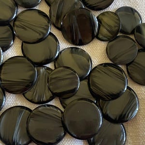Puede incluir: Un conjunto de botones redondos negros brillantes con un patrón arremolinado. Los botones, de diferentes tamaños, están dispersos sobre una superficie de tela clara. Estos botones son ideales para coser y proyectos de manualidades.