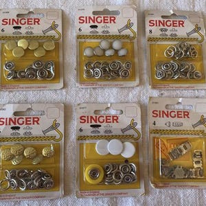 Pode incluir: São exibidas seis embalagens de materiais de costura da marca Singer. As embalagens contêm vários tipos de molas e fechos metálicos. A embalagem é amarela e branca com o logótipo Singer.