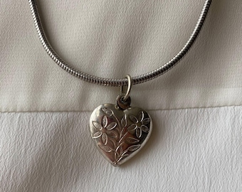 May include: A silver-toned necklace featuring a heart-shaped pendant. The pendant is adorned with an embossed floral design. The chain is a textured, snake-style chain, and the necklace is displayed against a white background.