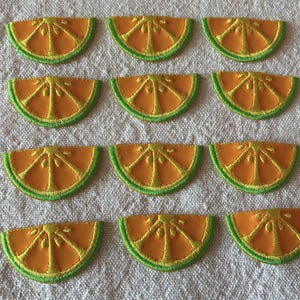 Puede incluir: Una colección de once parches de rodajas de naranja con detalles bordados en verde y amarillo. Los parches están dispuestos sobre una superficie texturizada de color blanco roto. Cada parche tiene forma de semicírculo, que se asemeja a una rodaja de fruta cítrica.