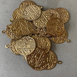 Pode incluir: Uma pilha de amuletos redondos de metal dourado com intrincados desenhos em relevo. Cada amuleto tem um pequeno laço na parte superior para fixar em joias ou outros trabalhos manuais. Os amuletos têm uma aparência vintage ou antiga.