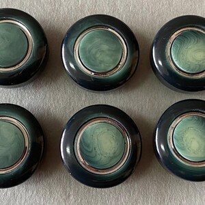 May include: Six circular buttons in dark teal and silver. Each button features a dark teal outer ring, a silver inner ring, and a lighter teal centre with a swirled pattern. These buttons are suitable for sewing or craft projects.