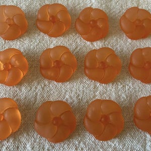 May include: Twelve orange, flower-shaped buttons with a frosted appearance are arranged on a textured, off-white surface. Each button has a central, raised detail and petal-like swirls. The buttons are likely for sewing or crafting.