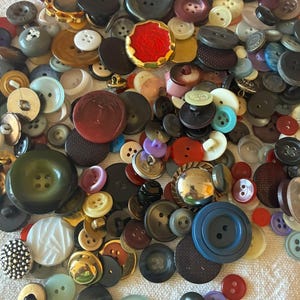 Pode incluir: Uma grande coleção de botões variados de diferentes formas, tamanhos e cores, incluindo vermelho, azul, verde, preto, branco e acabamentos metálicos. Os botões apresentam diversas texturas, padrões e números de furos, espalhados num monte denso.