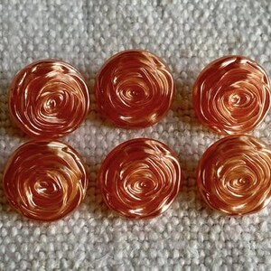 Pode incluir: Seis botões redondos cor de cobre com um design de rosa em espiral. Os botões estão dispostos em duas filas de três sobre uma superfície texturizada esbranquiçada. Os botões têm um acabamento metálico brilhante.