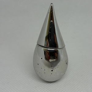 Może przedstawiać: Srebrny pojemnik w kształcie łzy z stożkową pokrywką. Pojemnik ma gładką, odblaskową powierzchnię z drobnymi niedoskonałościami. Pokrywka jest również srebrna i zwęża się do ostrego punktu. Przedmiot wydaje się być przedmiotem dekoracyjnym lub funkcjonalnym.