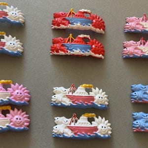 May include: A selection of miniature boat ornaments in various colours. Each boat has a red, white, and yellow design, set against blue and white waves, and is flanked by smiling suns or sea creatures. Likely magnets or small decorative items.
