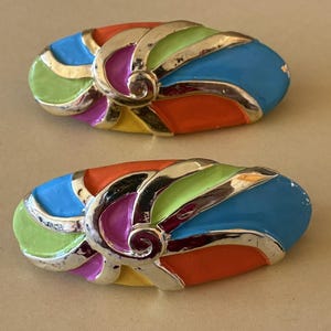 Pode incluir: Duas broches ovais vintage e coloridas. Cada broche apresenta uma moldura de metal prateado com secções de esmalte azul, verde, laranja, roxo e amarelo. O design inclui um detalhe em espiral.