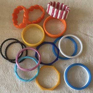 May include: A collection of colorful bracelets in various styles and designs. The bracelets include orange, yellow, pink, blue, white, and black. Some are solid, while others have geometric shapes or striped patterns. The bracelets are made of plastic.