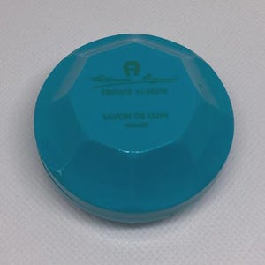 May include: A turquoise, faceted, round container with the text "Aigner Private Number SAVON DE LUXE" in green. The container has a geometric design and a shiny finish.