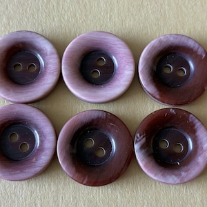 Pode incluir: Seis botões grandes e redondos com dois furos cada. Os botões têm uma aparência marmorizada, com tons de castanho e malva. Os botões estão dispostos em duas filas de três.