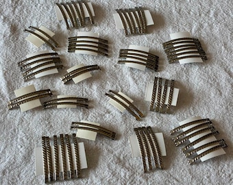 May include: A collection of metallic hair clips featuring a twisted rope-like texture. Each clip is attached to a small white rectangular card. The clips are finished in a muted bronze or antique gold tone and are arranged in various groupings.