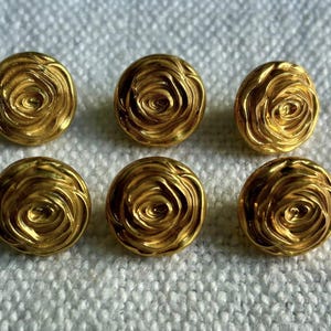 Puede incluir: Seis botones dorados con diseño de rosa. Los botones son redondos con un patrón en espiral en el centro, que se asemeja a una rosa. Los botones están sobre una superficie blanca texturizada.