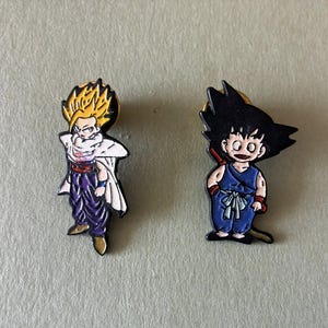 May include: Two enamel pins featuring anime characters. The left pin depicts a figure with spiky yellow hair, a white cape, and purple pants. The right pin shows a figure with black hair, a blue sleeveless top, and blue pants with a white sash.