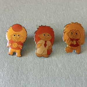 Puede incluir: Tres pines de esmalte con personajes de dibujos animados de pelo alborotado. El pin de la izquierda muestra un personaje con camiseta naranja y un smiley. El central muestra un personaje con camisa blanca y corbata roja. El de la derecha lleva una prenda roja.
