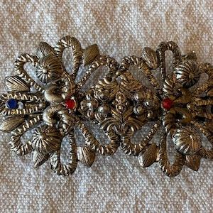 Pode incluir: Um gancho de cabelo em metal dourado com um design simétrico de folhas texturizadas e formas florais arredondadas. O acessório é decorado com dois pequenos strass azuis nas bordas externas e dois pequenos strass vermelhos perto do centro.