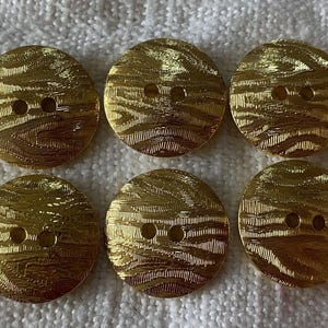 May include: Six gold-coloured, round buttons with a textured, wave-like pattern. Each button has two holes for sewing. The buttons are arranged in two rows of three.