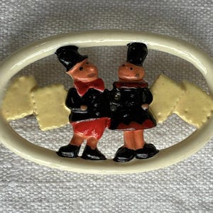 Può includere: Spilla ovale in ceramica con due figure in cappotti e cappelli neri, con dettagli rossi e neri. Le figure sono affiancate in una cornice color crema, con forme gialle su entrambi i lati. La spilla è probabilmente vintage.