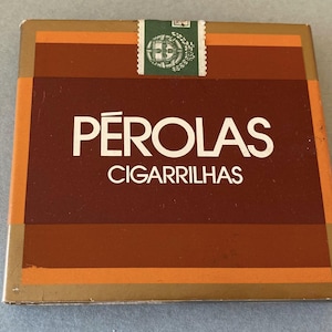 May include: A rectangular box of "Pérolas Cigarrilhas" cigarettes. The box has a brown background with orange and gold accents. The text "PÉROLAS" is in large white letters, with "CIGARRILHAS" below. A green stamp is at the top.