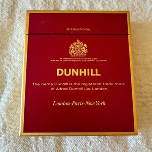 May include: A rectangular Dunhill cigarette box in deep red with gold trim and lettering. The front features the text International, the royal coat of arms, and the inscription By Appointment to Her Majesty the Queen, Suppliers of Smokers Requisites Alfred Dunhill Ltd. London, followed by the brand name, trademark notice, and London Paris New York.