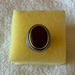 Op de afbeelding: Een zilveren ring met een ovaalvormige donkerrode steen in een goudkleurige zetting. De ringband heeft een gestructureerd, geribbeld ontwerp rondom de centrale steen, wat zorgt voor een gelaagd uiterlijk met contrasterende metalen afwerkingen.